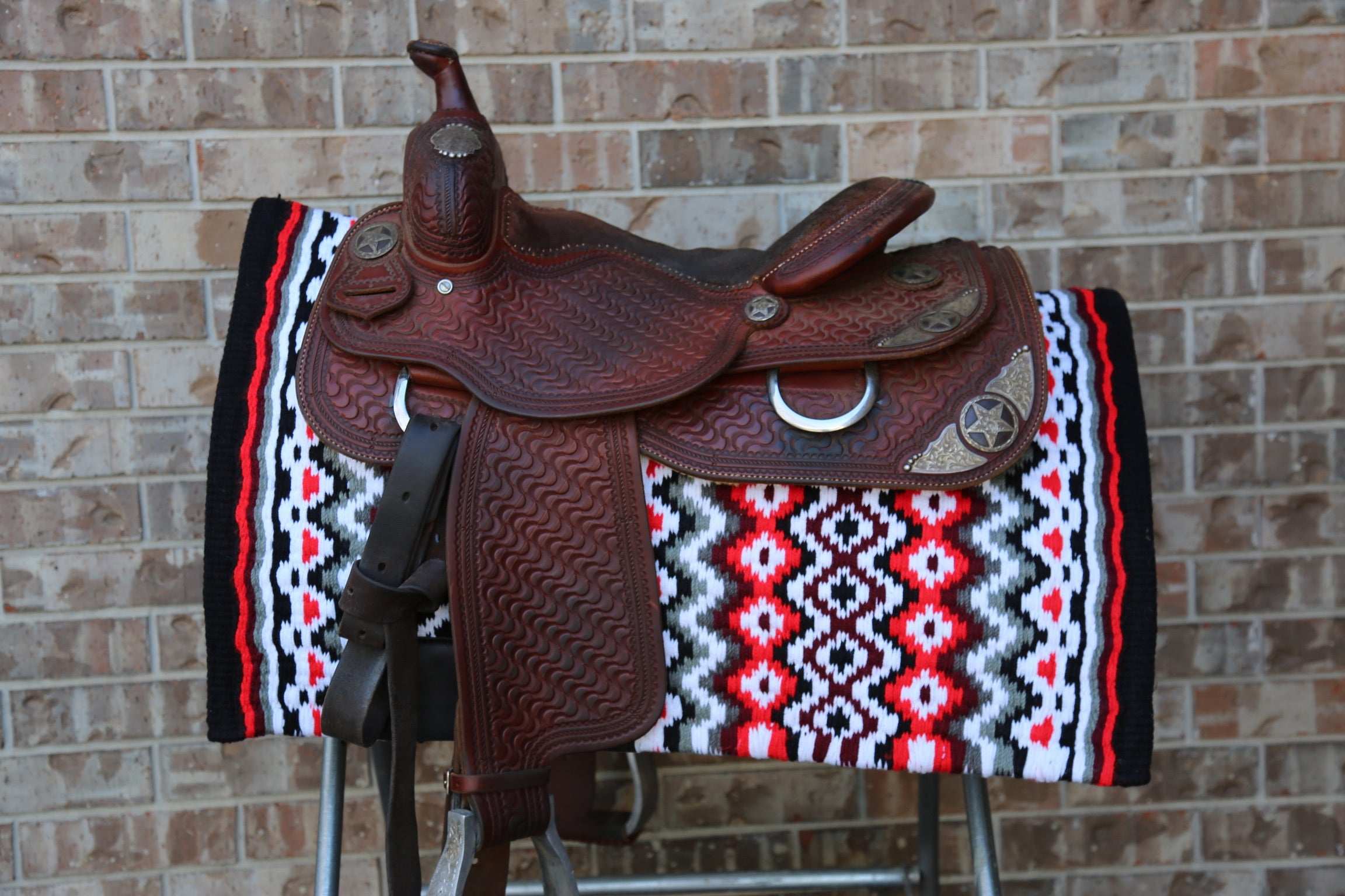 Custom Western Saddle Pad in Red, Brown, White, Black geometric color pattern| Limited Edition displayed on saddle rack and western saddle on top with brick background
