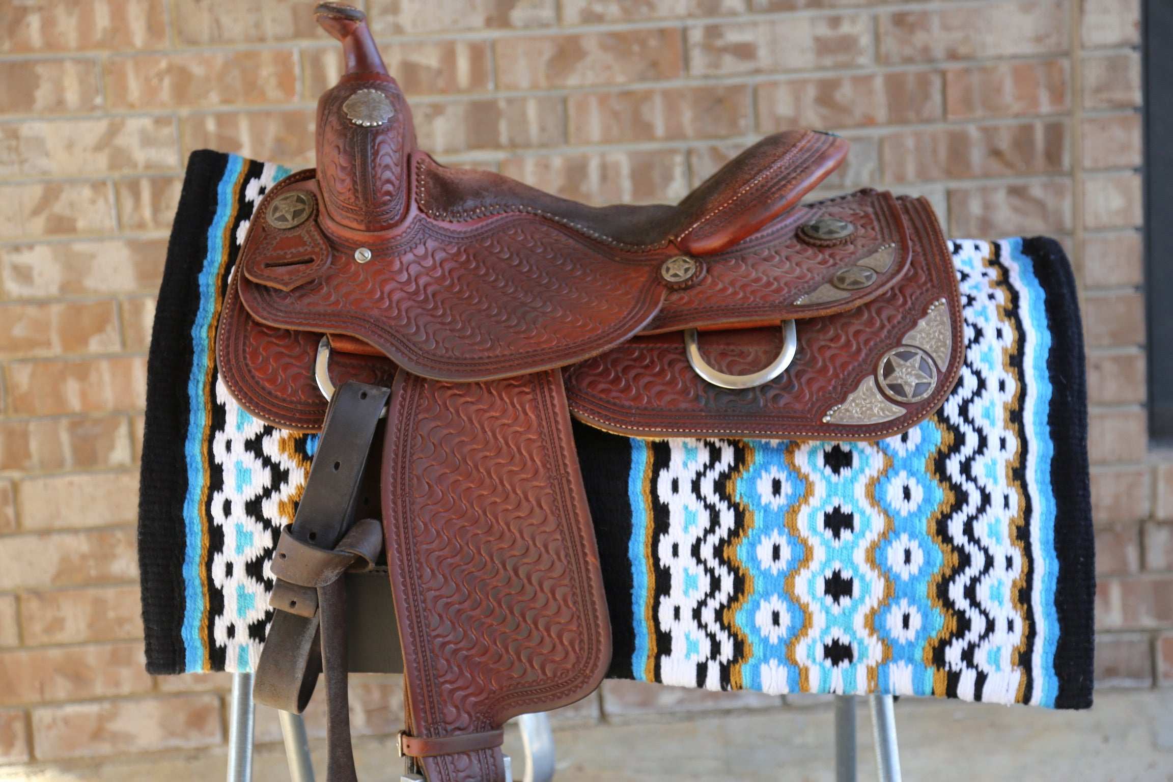 Custom Western Saddle Pad in Blue, Gold, White, Black geometric colors pattern on top of a saddle rack  and western saddle on top with brick wall background | Limited Edition