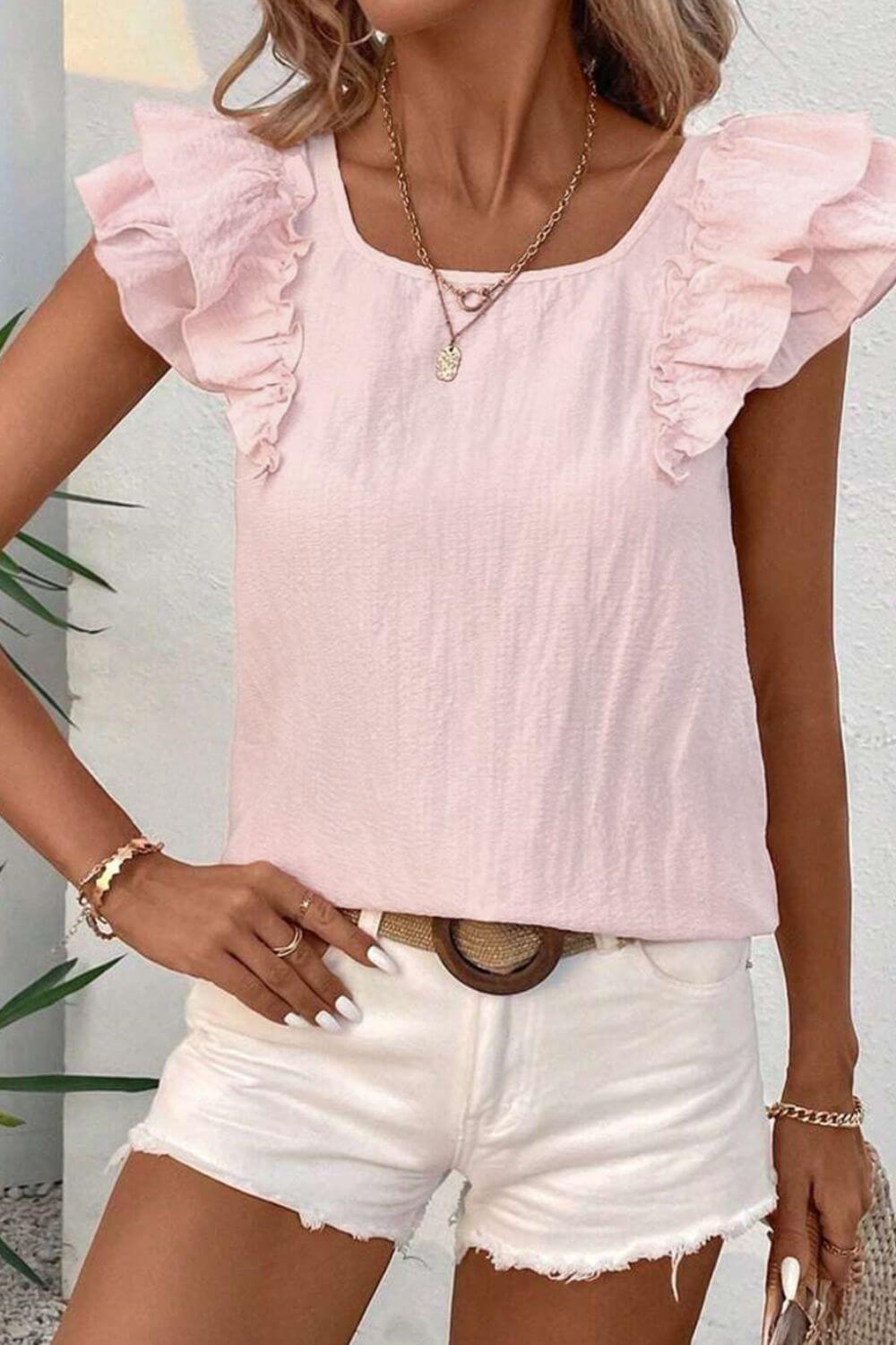 Woman wearing a pink top with ruffled sleeves and white shorts, standing against a neutral background.